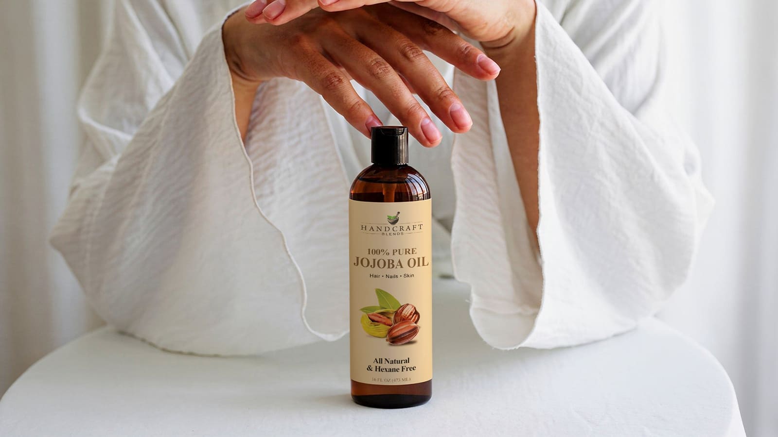 100% pure jojoba oil bottle for hair, nails, and skin on white background with hands above