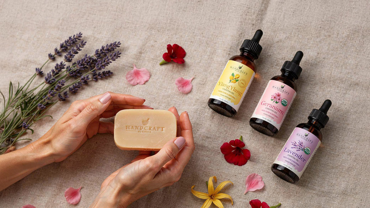 Handmade floral soap bars beside bottles of lavender, geranium, and ylang ylang essential oil