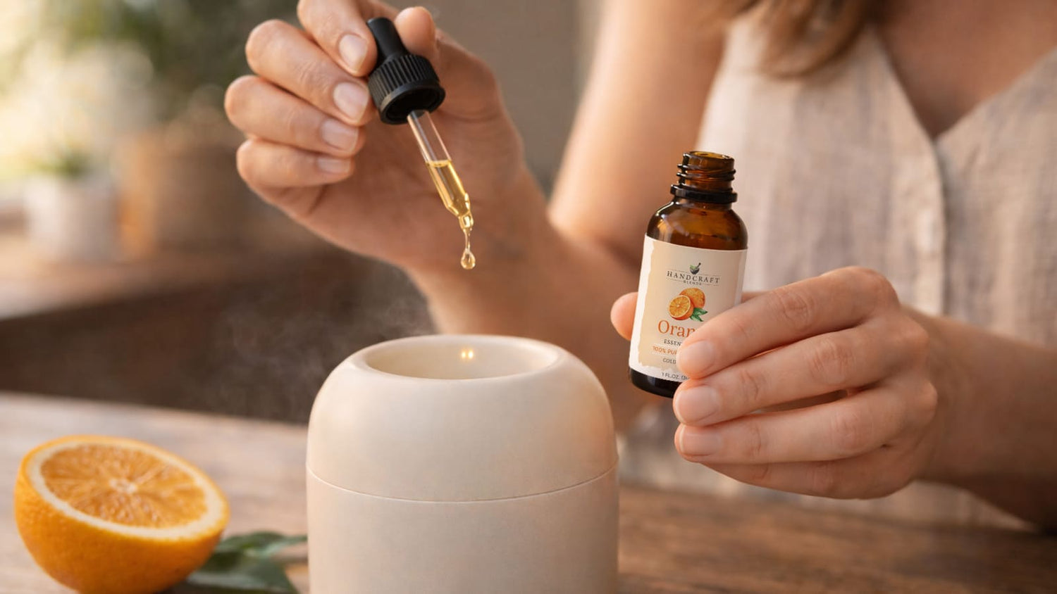Adding orange essential oil with dropper to a diffuser, fresh orange and leaves nearby