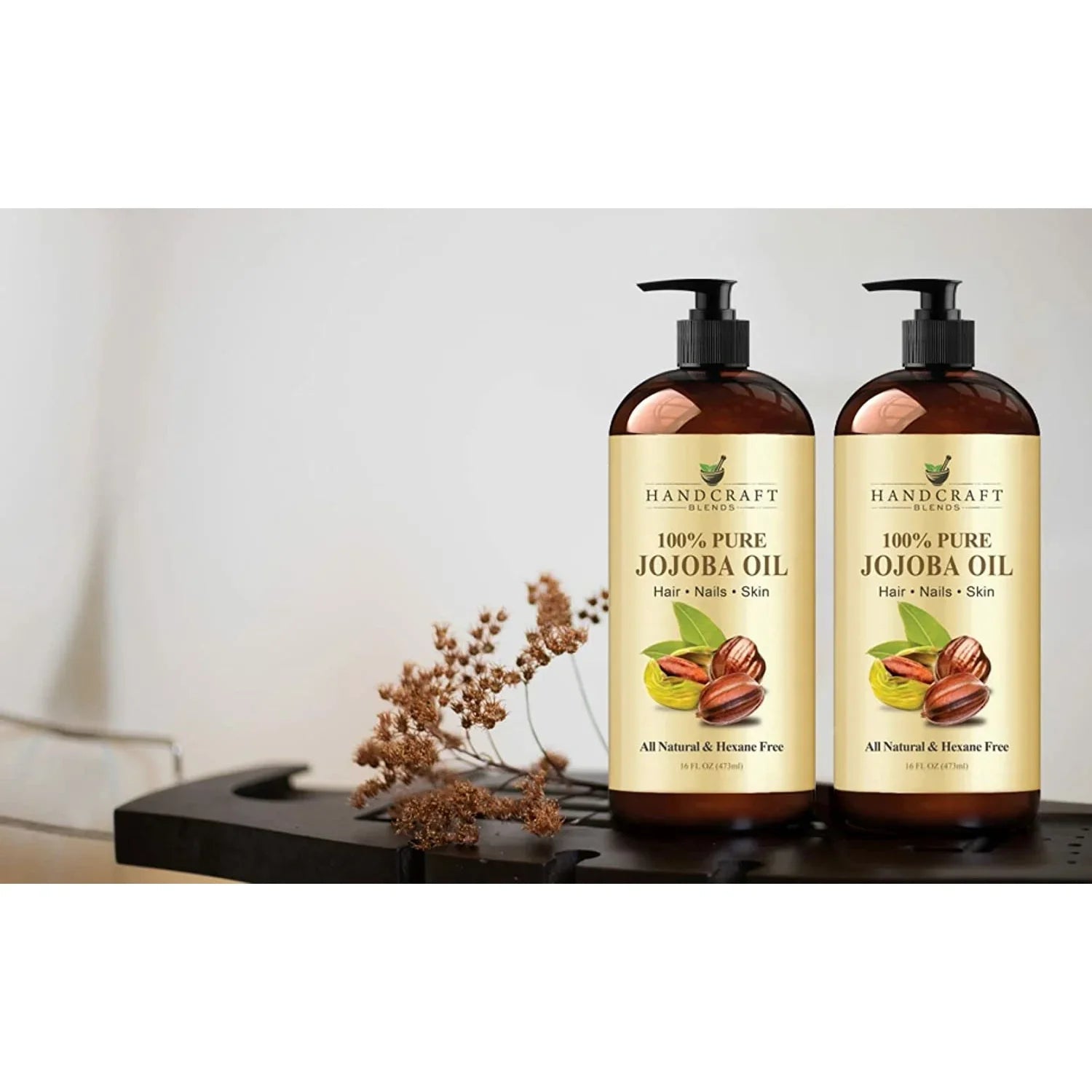 Two bottles of 100% pure jojoba oil for hair, nails, and skin on a table with dried flowers.